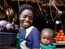 photo of smiling woman carrying bucket of produce with baby on her back